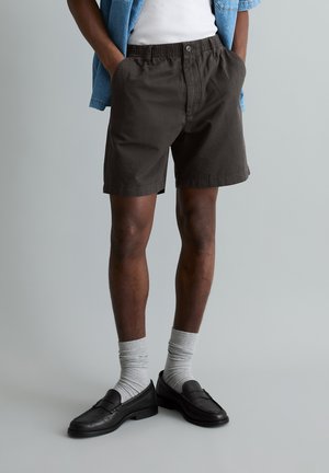 Person wearing black casual shorts, white socks, black loafers, a white tank top, and a blue short-sleeve shirt with hands in pockets.