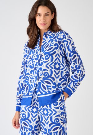 Mujer con camisa y pantalones de manga larga con un patrón ornamentado azul y blanco a juego, de pie con una mano en el bolsillo, sobre un fondo liso.