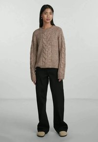 Gestrickter Pulli aus weichem Beige, mit strukturierten Mustern und lockeren Ärmeln, kombiniert mit weiten schwarzen Hosen und hellen Schuhen.