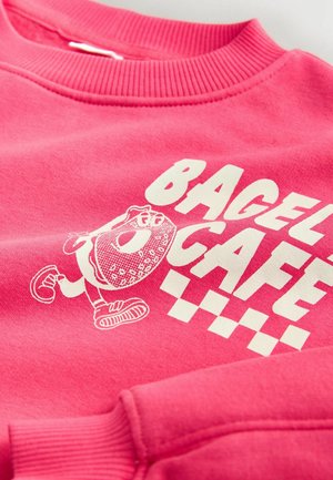 Rožinis džemperis su balta bagelio grafika ir užrašu „BAGEL CAFE“ su languotais akcentais. Minkštas audinys su ribotu apykakle.