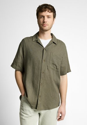 Jeune homme aux cheveux bouclés portant une chemise boutonnée à manches courtes vert olive sur un t-shirt blanc, debout avec une main dans la poche.