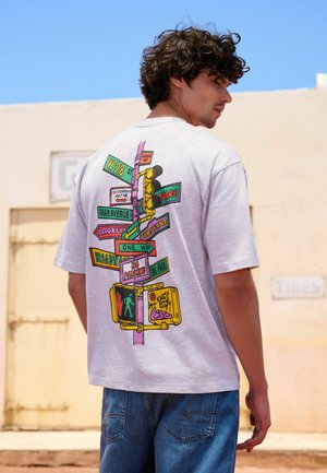 Young man with curly hair wearing a white T-shirt featuring colorful street signs and traffic lights graphic on the back, standing outdoors.