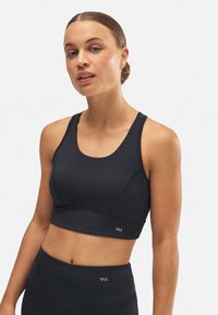 Sostén deportivo negro con tirantes anchos, tejido suave y diseño ajustado. Presenta un pequeño logo en el lado izquierdo y un escote redondo simple.