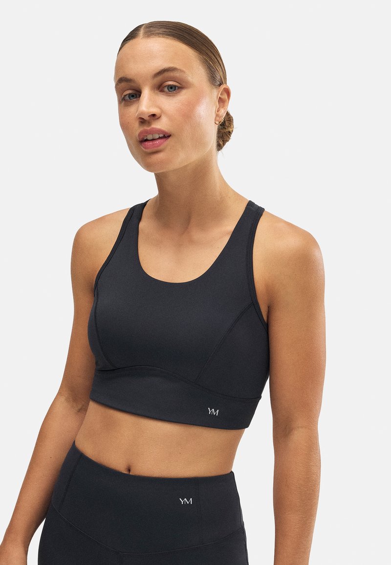 Sostén deportivo negro con tirantes anchos, tejido suave y diseño ajustado. Presenta un pequeño logo en el lado izquierdo y un escote redondo simple.