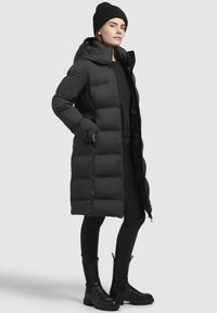 Giacca a vento nera con cappuccio, di lunghezza lunga, con un motivo trapuntato. Indossata sopra un top nero e leggings, abbinata a stivaletti neri.