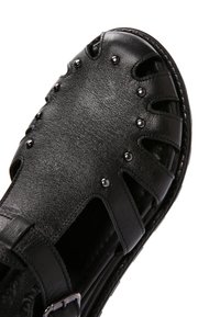 Sandalo aperto in pelle nera con borchie metalliche, design traforato, suola cucita e cinturino alla caviglia regolabile con fibbia metallica.