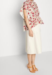 Donna che indossa una blusa a cappotto floreale beige, una gonna midi color crema e sandali con tacco arancioni, in piedi su uno sfondo bianco.