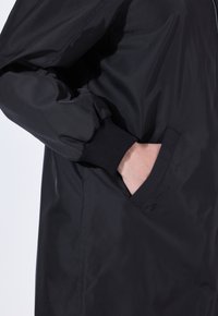 Cazadora negra con un tejido liso y brillante, puños acanalados y bolsillos laterales. El diseño incluye un corte holgado y costuras mínimas.