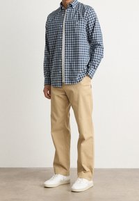 Mężczyzna w niebiesko-białej koszuli w kratę na guziki, na białym t-shircie, beżowe spodnie i białe trampki stojący na szarej podłodze.