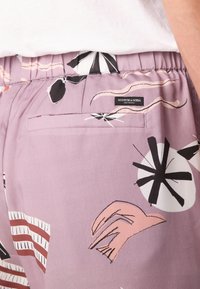 Shorts violets clairs avec des motifs abstraits noirs, blancs et roses, taille élastique et une poche arrière unique arborant une étiquette de marque.