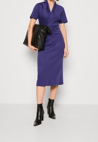 Fialové šaty ke kolenům s krátkými rukávy a ozdobným přetočením vpředu. Kombinovány s velkým černým psaníčkem a černými kotníkovými botami na podpatku.
