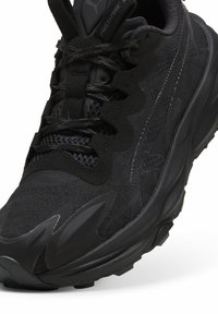 Chaussure de sport noire avec une tige en maille texturée, un design superposé, un col rembourré et une semelle en caoutchouc présentant des motifs de bande de roulement proéminents.