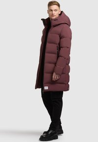 Maroon puffer coat with a hood, featuring a zip front, side pockets, and a quilted design. Worn over a black outfit with black shoes.