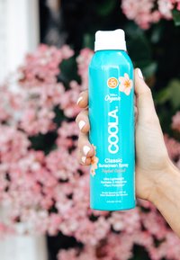 Turkusowa butelka sprayu z białą zakrętką, oznaczona napisem "COOLA Classic Sunscreen Spray", z kwiatowymi akcentami i szczegółami SPF 30.