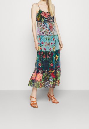Kobieta ubrana w bezrękawnikową, wielokolorową sukienkę maxi z kwiatowym wzorem patchwork, z cienkimi ramiączkami i pomarańczowymi sandałami na obcasie z paskami.