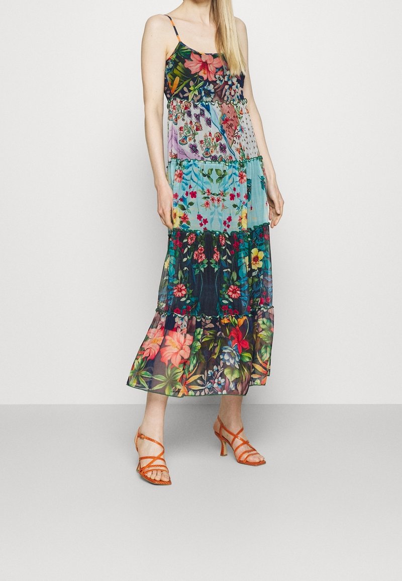 Femme portant une robe maxi patchwork florale multicolore sans manches avec fines bretelles et sandales à talons orange à brides.