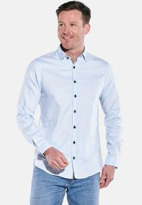 Mann in einem hellblauen, langärmeligen Hemd mit Knöpfen und blauen Jeans, der steht und lächelt, vor einem schlichten weißen Hintergrund.
