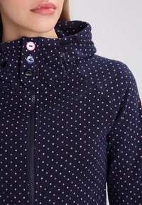 Hoodie marine avec des pois roses et blancs, doté d'une fermeture éclair à l'avant, d'une capuche avec cordon de serrage et de boutons décoratifs. Texture de tissu doux.