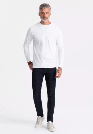 Weißes Langarmshirt, kombiniert mit dunklen Jeans und hellen Sneakern. Das Outfit hat einen klassischen Rundhalsausschnitt und ein schlankes Design.