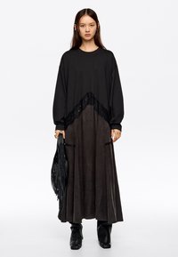 Czarna sukienka z długim rękawem, zdobiona koronkowymi akcentami i kontrastującymi teksturami, o zwiewnym kroju i dwóch bocznych kieszeniach. Modelka trzyma torebkę z frędzlami.