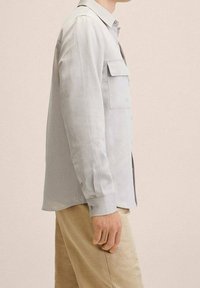 Camisa gris claro abotonada con mangas largas, dos bolsillos delanteros y corte recto. Tejido liso con un sutil brillo y mangas con puños.