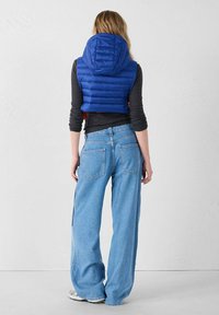 Chaleco azul acolchado con capucha sobre una camiseta de manga larga oscura, combinado con vaqueros de pierna ancha y color azul claro, con bolsillos traseros y un ajuste relajado.