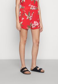 Tuta rossa con motivo floreale, vita elastica e design con pantaloni corti. Indossata con sandali neri con doppie cinghie e soletta in sughero.