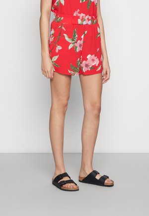 Combinaison rouge à motif floral, taille élastique et design short. Portée avec des sandales noires à doubles brides et semelle en liège.
