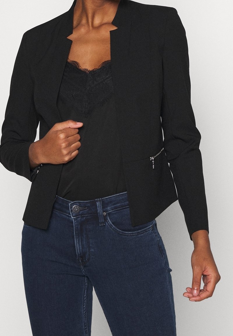 Femme portant un blazer noir ouvert avec des détails zippés sur un haut noir bordé de dentelle et un jean bleu foncé, tenant le revers du blazer d'une main.