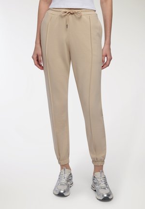 Pantaloni jogger beige con polsini elastici e vita con cordino, indossati con scarpe da ginnastica argento e bianche.