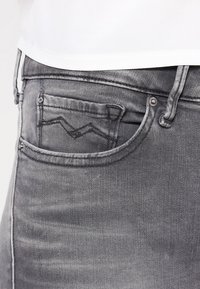 Jeans de mezclilla gris con un bolsillo trasero que presenta un diseño de puntada en zigzag. La tela tiene una textura suave con un aspecto ligeramente desgastado.