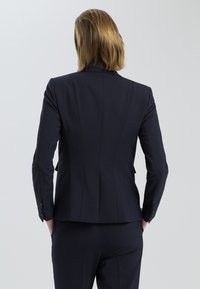 Navy fitted blazer with structured shoulders, back seams, and buttoned cuffs, made from smooth fabric. Pockets on both sides.