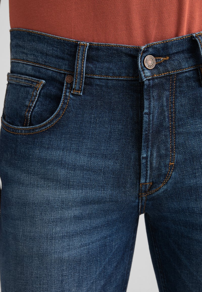 Tmavě modré džíny z denimu s viditelným prošitím, přední kapsou, poutky na pásek a knoflíkovým zapínáním, které jsou nošeny se topem v rezavé barvě.