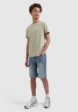 Tiener die een beige t-shirt, spijkershorts, witte sokken en zilveren sneakers draagt, staat met één hand in de zak en kijkt opzij.