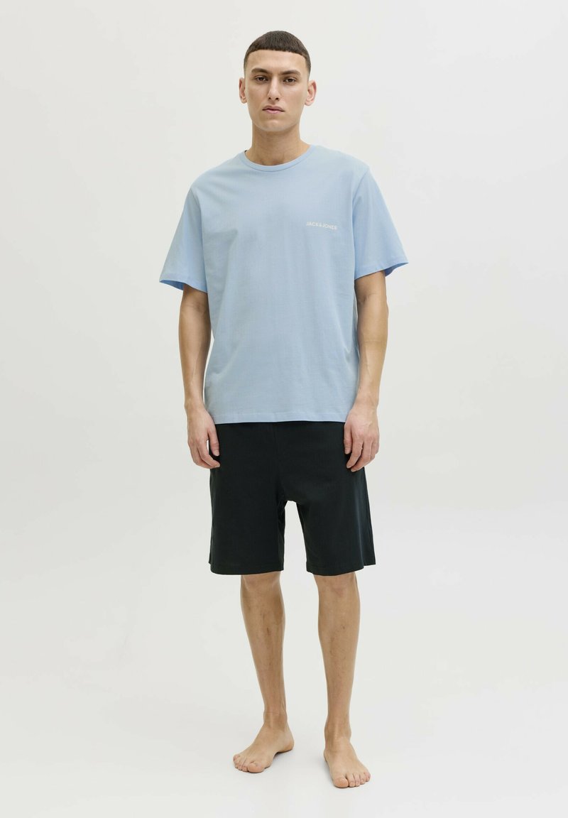 Junger Mann steht barfuß, trägt ein hellblaues kurzärmeliges T-Shirt und schwarze knielange Shorts vor einem schlichten weißen Hintergrund.