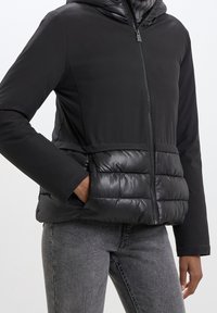 Giacca nera con zip e parte inferiore imbottita, indossata sopra jeans grigi, con una mano in tasca e l'altra rilassata di fianco.