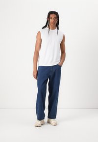 Camicetta bianca senza maniche abbinata a jeans denim blu a taglio largo e sneaker beige chiaro. Tessuto morbido, forma rilassata, design minimale.