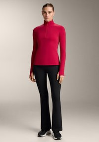 Figursyet langærmet top i lys pink med en halv lynlås, kombineret med sorte flared leggings og sorte sportssko på en neutral baggrund.