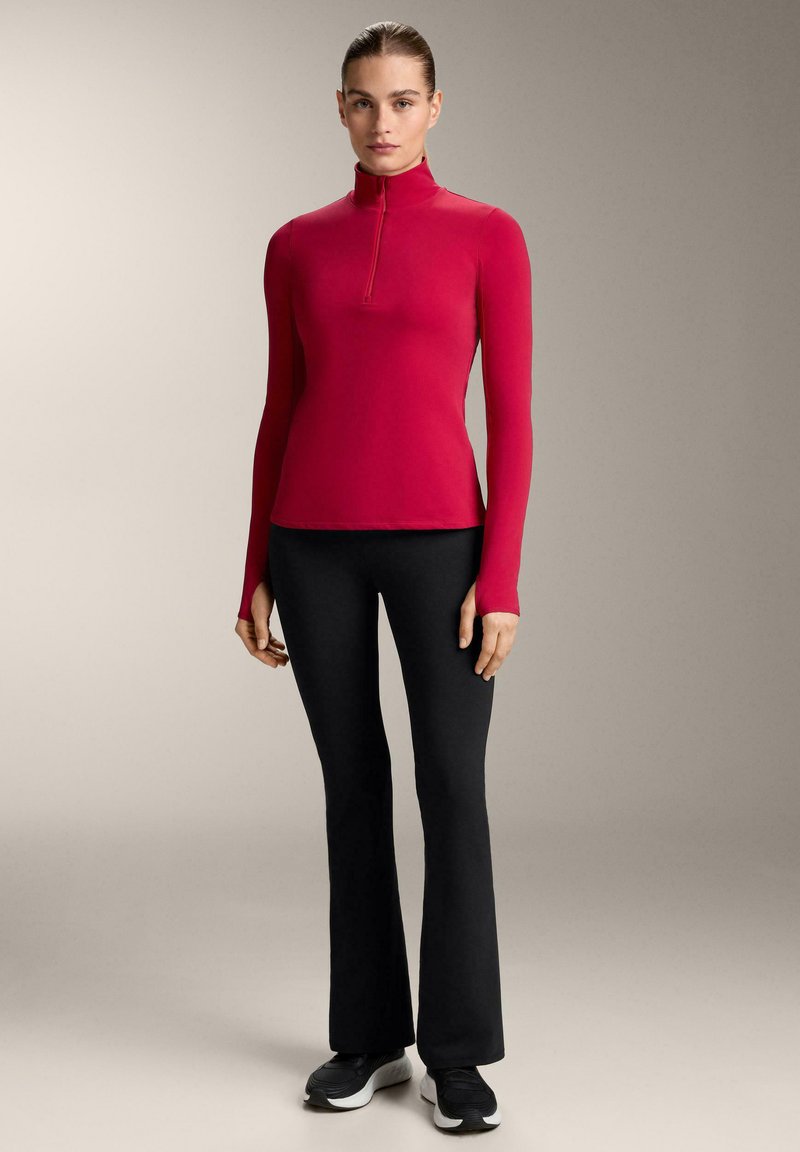 Figursyet langærmet top i lys pink med en halv lynlås, kombineret med sorte flared leggings og sorte sportssko på en neutral baggrund.