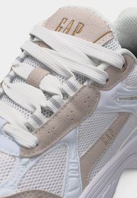Baskets GAP blanches et beiges avec des panneaux en maille et en daim, lacets blancs, et logo GAP sur la languette et le côté.