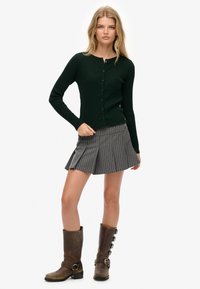 Ciemnozielony, prążkowany kardigan z długimi rękawami i zapięciem na guziki, w połączeniu z szarym spódnicą plisowaną z pionowymi paskami oraz brązowymi botkami sięgającymi do kolan.