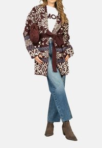 Cardigan lungo a fantasia bordeaux e crema con cintura da annodare, indossato su una camicia bianca, abbinato a jeans azzurri e stivaletti marroni.