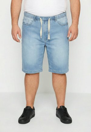 Mand iført lyseblå denimshorts med hvid snor, hvid skjorte og sorte sneakers, stående mod en ensfarvet baggrund.