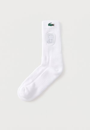 Egyetlen fehér csapatzokni zöld krokodil logóval és teniszütő emblémával a bordázott szegélyen, egyszínű háttér előtt.