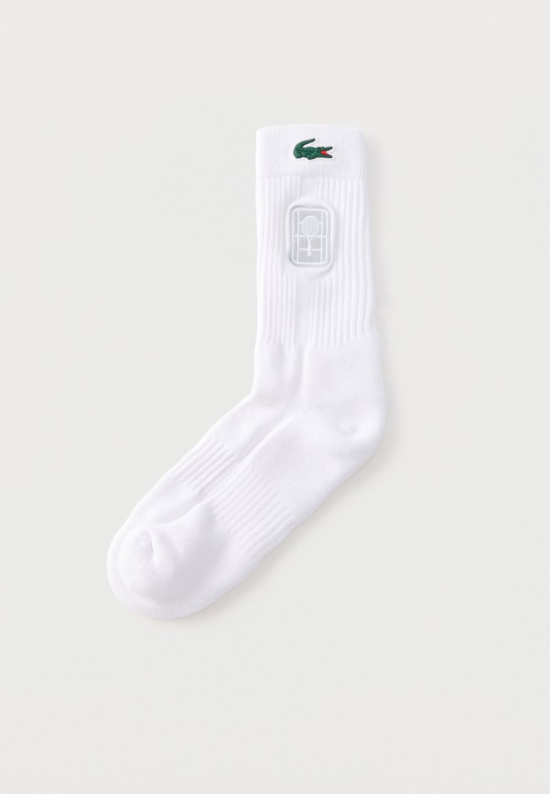 Μονό λευκό καλτσάκι ομάδας με πράσινο λογότυπο κροκόδειλου και έμβλημα ρακέτας τένις σε ραβδωτό μανσέτα σε απλό φόντο.