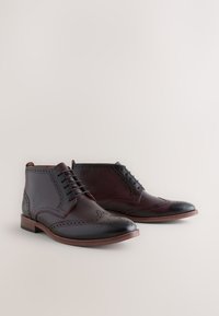 Braune Leder-Stiefeletten mit Brogue-Details, die ein glattes Finish, kontrastierende dunkle Akzente und runde Zehen aufweisen. Dezentes Schnürdesign.