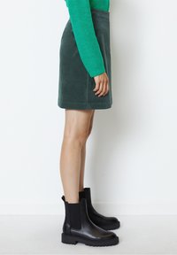 Grüner Wildleder-Minirock mit zwei seitlichen Taschen, kombiniert mit schwarzen Knöchelstiefeln mit elastischen Seitenteilen und robusten Sohlen.