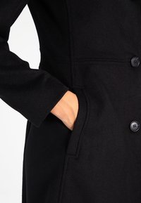 Svart ullblandad kappa med skräddarsydd design, knappar och sidofickor. Närbild av en hand i fickan och detalj av ärmen.