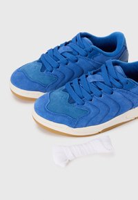 Des baskets en suède bleu présentent un design superposé avec des accents texturés et des lacets bleus assortis. Une dentelle blanche se trouve à côté des chaussures.