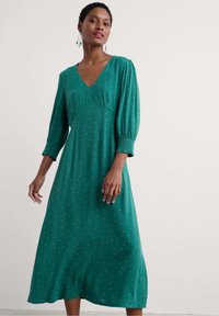 Vestido verde de manga larga con escote en V, que presenta una silueta fluida, puños fruncidos y pequeños puntos blancos dispersos por toda la prenda.